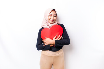 A happy young Asian Muslim woman wearing a hijab feels romantic shapes heart gesture expressing tender feelings and holding a red heart-shaped paper