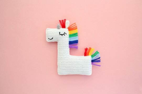 Crochet amigurumi handmade stuffed soft white unicorn toy with rainbow mane on pink background. Handwork, hobby. Craft diy newborn pregnancy concept. Knitted doll for little baby. Close up flat lay