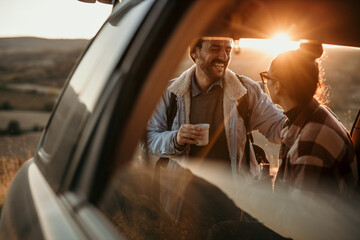 Young couple talking, drinking a coffee watching the sunset from their car. Couple travel concept