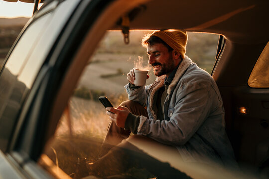 A handsome man in a denim jacket is sitting in the car truck, browsing a phone and drinking a hot drink in nature during the sunset.