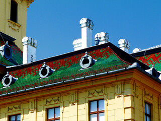 colorful steep ceramic tile mansard roof. red and green enamel finish. decorative white stucco chimneys. old classic apartment building detail. wood windows. blue sky. yellow stucco residential facade