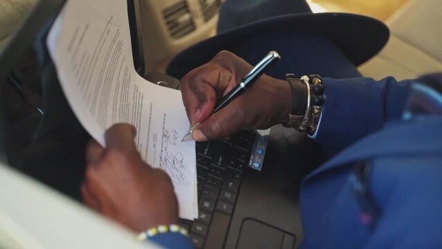African businessman is signing a document in the back of a car, having laptop top and writing on paper on top of it.