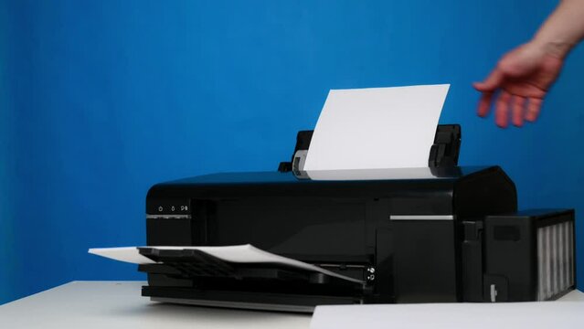 A hand inserts blank sheets of A4 paper into the printer on a blue background.Office, printing equipment.