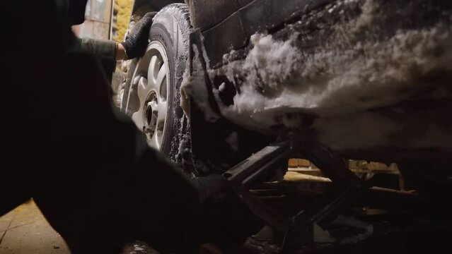 Car mechanic picks up car on doncrat in old dirty garage. On carbolic spine or lot of snow stuck to body and arches with wheel. Wheel removal and suspension repair. Cold or winter.