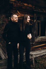 Fototapeta na wymiar a man and a woman in black clothes near an old wooden house. photo in dark brown tones. old abandoned house and cloudy cold weather. beautiful stylish couple on the background of a brown wooden wall.