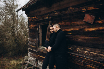 a man and a woman in black clothes near an old wooden house. photo in dark brown tones. old abandoned house and cloudy cold weather. beautiful stylish couple on the background of a brown wooden wall.