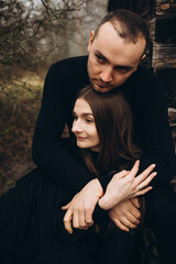 a man and a woman in black clothes sit near an old wooden house. photo in dark brown tones. old abandoned house and cloudy cold weather. hug a loved one