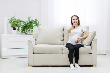Cute pregnant woman sitting on sofa with remote control.