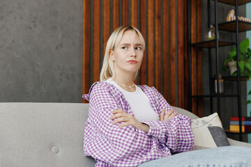 Young sad woman wear casual clothes look aside hold hands crossed folded sits on grey sofa couch stay at home hotel flat rest relax spend free spare time in living room indoor. People lounge concept.