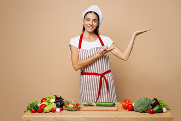 Young housewife housekeeper chef cook latin woman wear apron toque chefs hat work at table kitchenware point hands arms aside area isolated on plain pastel light beige background Process cooking food