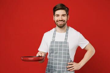 Young smiling confident male housewife housekeeper chef cook baker man wear grey apron hold in hand frying pan stand akimbo arm on waist isolated on plain red background studio. Cooking food concept.