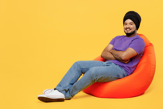 Full body fun devotee Sikh Indian man ties his traditional turban dastar wear purple t-shirt sit in bag chair hold hands crossed folded look camera isolated on plain yellow background studio portrait.