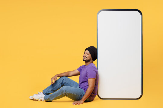 Full body fun devotee Sikh Indian man ties his traditional turban dastar wear purple t-shirt sit near big huge blank screen area mobile cell phone isolated on plain yellow background studio portrait.