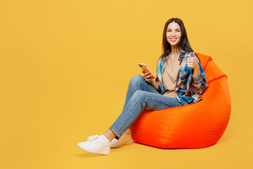 Full body young fun woman wear blue shirt beige t-shirt sit in bag chair hold in hand use mobile cell phone show thumb up isolated on plain yellow background studio portrait. People lifestyle concept.