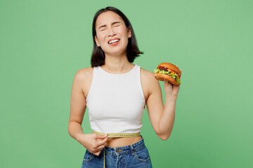 Young crying sad woman wearing white clothes holding measuring tape on waist eating burger isolated...