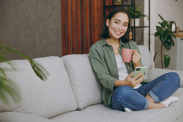 Full body young woman of Asian ethnicity wearing casual clothes hold use mobile cell phone drink coffee sit on grey sofa couch stay at home hotel flat rest relax spend free spare time in living room.