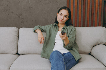 Fototapeta premium Young bored sad un happy woman of Asian ethnicity wears casual clothes watch TV switch channels sits on grey sofa couch stay at home hotel flat rest relax spend free spare time in living room indoor.