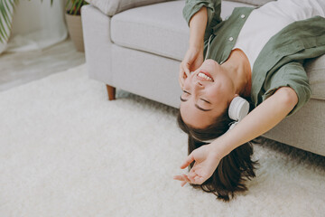 Young fun woman of Asian ethnicity wearcasual clothes headphones listen to music close eyes lay down on grey sofa couch stay at home hotel flat rest relax spend free spare time in living room indoor.
