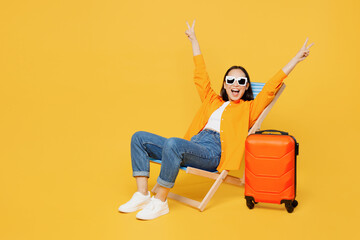 Young woman wear summer clothes sit in deckchair spread hands show v-sign isolated on plain yellow background. Tourist travel abroad in free spare time rest getaway. Air flight trip journey concept.