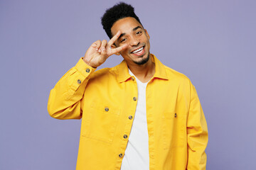 Young happy man of African American ethnicity wear yellow shirt t-shirt showing cover eye with victory sign isolated on plain pastel light purple background studio portrait. People lifestyle concept.