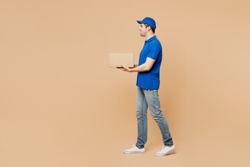 Naklejka na ściany i meble Full body side profile view delivery guy employee man wear blue cap t-shirt uniform workwear work as dealer courier hold cardboard box go walk isolated on plain light beige background Service concept