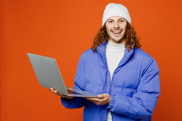 Young student smiling happy IT man with long curly hair wear hat purple ski padded jacket casual clothes hold use work on laptop pc computer isolated on plain orange red background studio portrait.