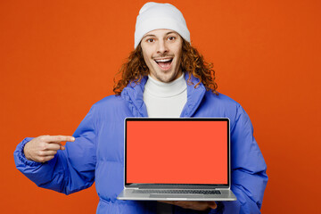 Young IT man with long curly hair wear hat purple ski padded jacket casual clothes hold use work point on laptop pc computer with blank screen workspace area isolated on plain orange red background.