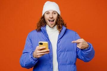 Young fun surprised man with long curly hair wear hat purple ski padded jacket casual clothes hold use point index finger on mobile cell phone isolated on plain orange red background studio portrait. © ViDi Studio