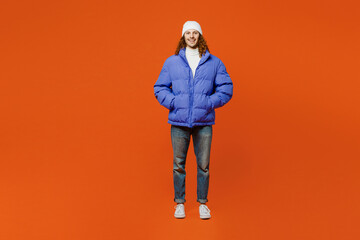 Full body smiling happy fun young man with long curly hair wear hat purple ski padded jacket casual clothes hold hands in pockets look camera isolated on plain orange red background studio portrait. © ViDi Studio
