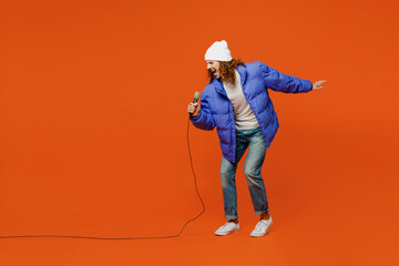 Full body expressive young man with long curly hair wears hat purple ski padded jacket casual clothes sing song in microphone at karaoke club isolated on plain orange red background studio portrait.