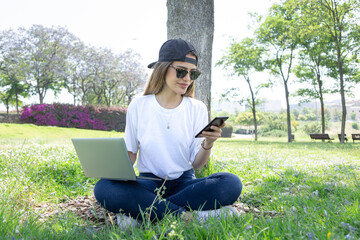 Front view of a happy student girl working with a laptop and smartphone in a green park of an university campus