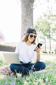 Front view of a happy student girl working with a laptop and smartphone in a green park of an university campus