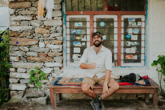 Smiling male hiker resting on bench with beverage in hand in mountains