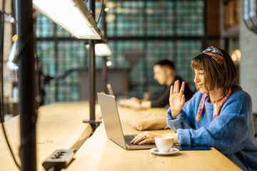 Young stylish woman works on laptop while sitting by shared table with a coffee drink at modern cafe. Concept of remote work from public place, digital freelance and modern lifestyle