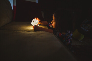 A little girl sits in the dark with her face lit up by a small lamp