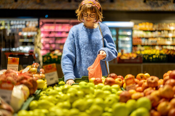 Young woman chooses apples to buy and puts them into mesh bag at fruit department of a supermarket. Concept of consumerism of healthy food and use of reusable bags