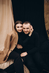 Fototapeta na wymiar a man and a girl in black clothes and a beige coat on a black and beige fabric background. man and woman hugging, cold cloudy weather and dry yellow tall grass . stylish fashion portrait.