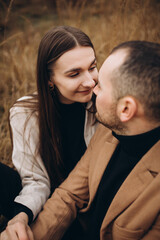 a man and a woman wearing black clothes and a beige coat are sitting and hugging in the tall dry grass. cloudy cold autumn weather. a stylish couple can