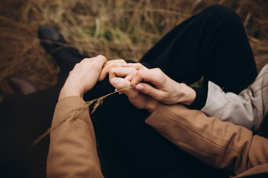 a man and a woman wearing black clothes and a beige coat are sitting and hugging in the tall dry grass. cloudy cold autumn weather. a stylish couple can