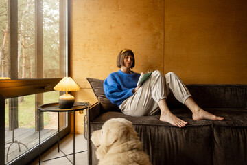 Woman reads a book while sitting relaxed on a couch by the window with her dog at wooden cottage in...