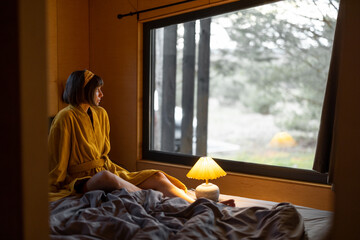 Woman in yellow bathrobe wakes up in tiny bedroom of a wooden cabin in forest. Good morning and rest in house on nature concept