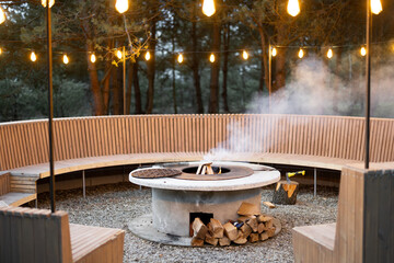 Beautiful lounge area with fireplace, round bench and illuminated garlands in forest at dusk