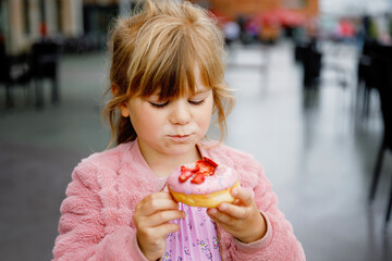 Happy little preschool girl eating sweet donut indoor. Blond child in the city. Cute kid and...