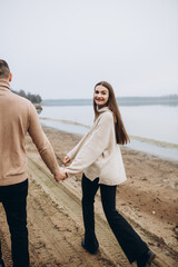a man and a woman are walking along the shore of the lake, cold foggy weather. romantic photo in beige tones. holding a woman's hand, a love story. beige sweaters. romantic photos by the lake