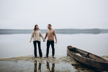 a man and a woman stand on the shore of a lake, cold foggy weather. A wooden boat. holding a woman's hand, a love story. beige sweaters. romantic photos against the background of a foggy lake