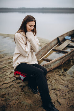 beautiful woman on the shore of a foggy lake, cold weather. A wooden boat on which a woman sits. a girl in a warm beige sweater, romantic photos against the background of a foggy lake