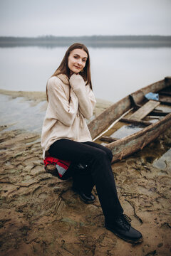 beautiful woman on the shore of a foggy lake, cold weather. A wooden boat on which a woman sits. a girl in a warm beige sweater, romantic photos against the background of a foggy lake