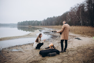 a man on the shore of a lake near a fire plays the guitar for a girl. foggy cold weather. beige men's coat. bonfire on the shore of the reservoir. photo in beige tones. romantic photo with a guitar 