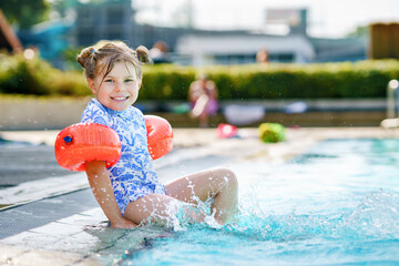 Little preschool girl with protective swimmies playing in outdoor swimming pool by sunset. Child learning to swim in outdoor pool, splashing with water, laughing and having fun. Family vacations.