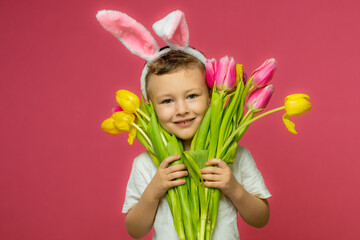 Obraz na płótnie Canvas Happy Easter. A child in a rabbit costume holds a bouquet of yellow and pink tulips on pink background. A charming baby with funny bunny ears. Spring Easter holiday. The concept of a happy childhood.